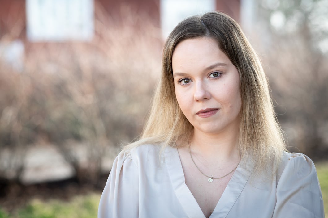 Alice Storm med långt hår har på sig en ljus blus och står utomhus, tittar mot kameran. I bakgrunden syns träd och en byggnad, som är suddiga.
