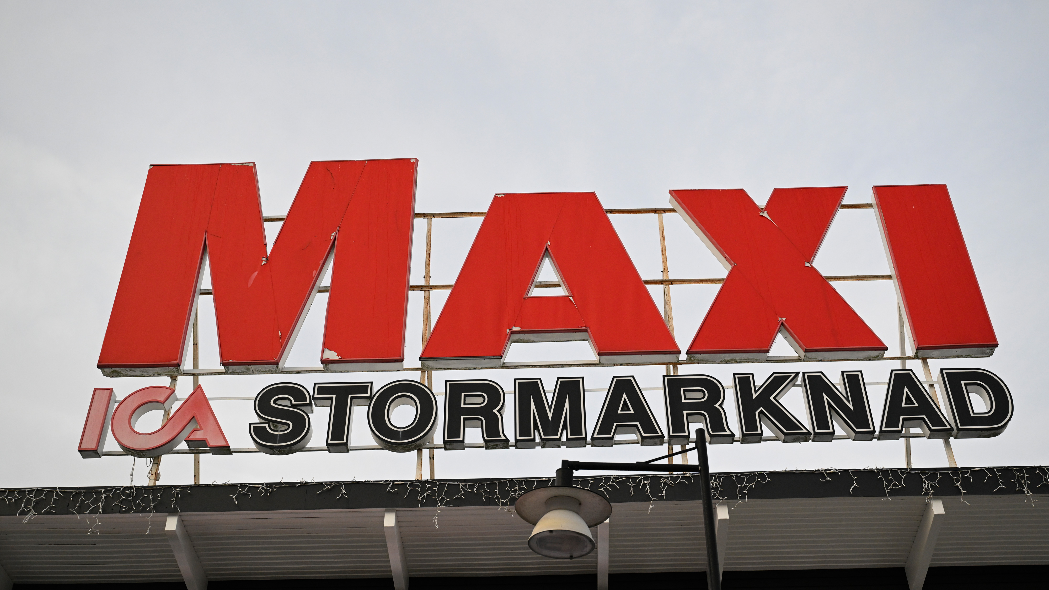 Stor skylt med texten "MAXI ICA Stormarknad" i rött och vitt på en byggnadsfasad mot molnig himmel.