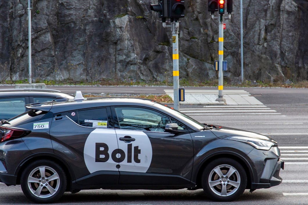 En bil med Bolt-logga står vid rött ljus på en stadsgata, med en bergssida i bakgrunden.