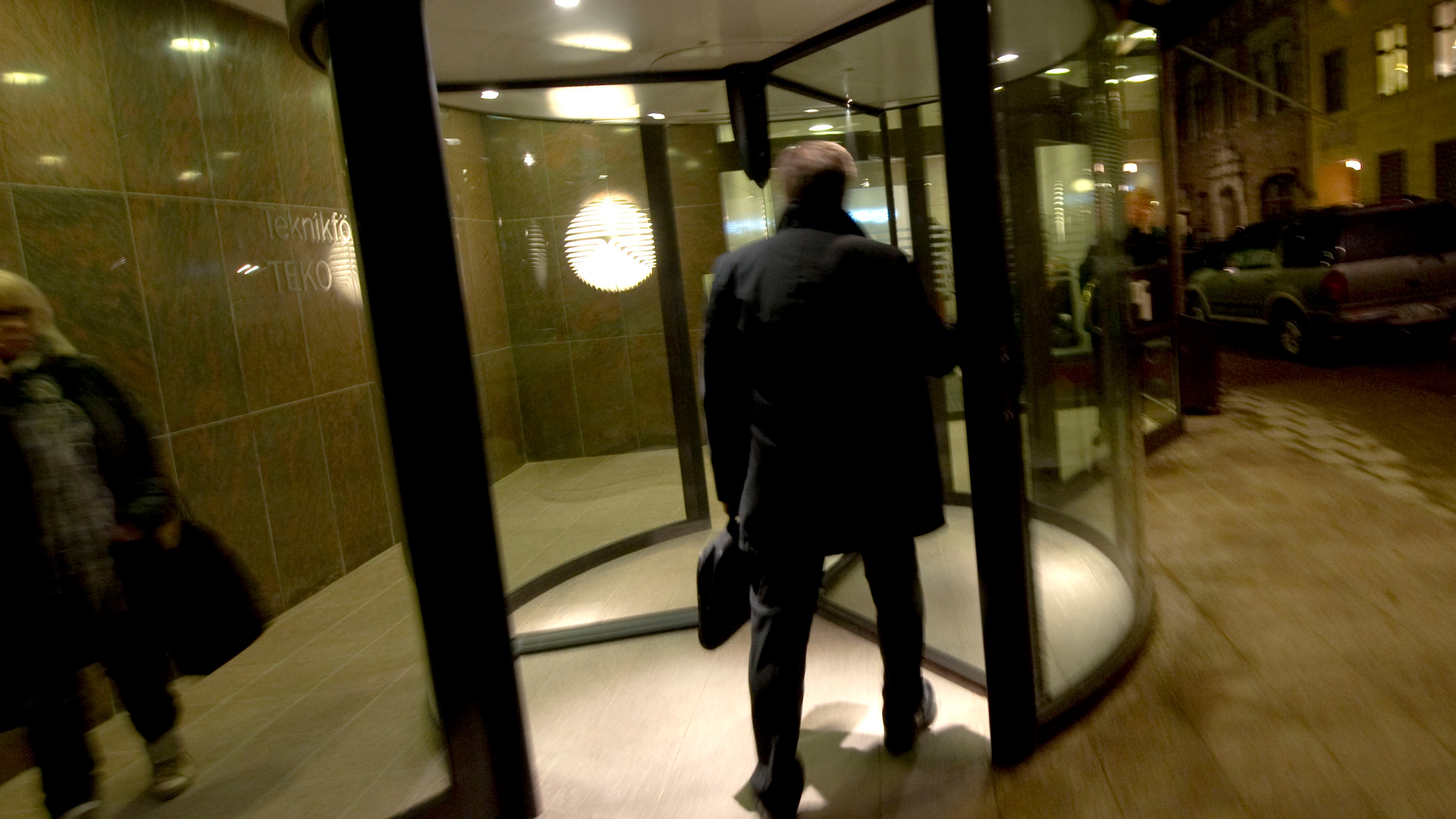 En person i en mörk rock går genom en svängdörr på kvällen och håller i en portfölj. En annan person lämnar platsen till vänster. I bakgrunden syns en svagt upplyst gata med parkerade bilar.