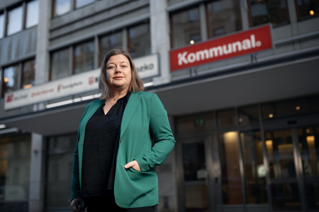 En person i en grön blazer står framför en byggnad med skyltar som visar ordet "Kommunal".