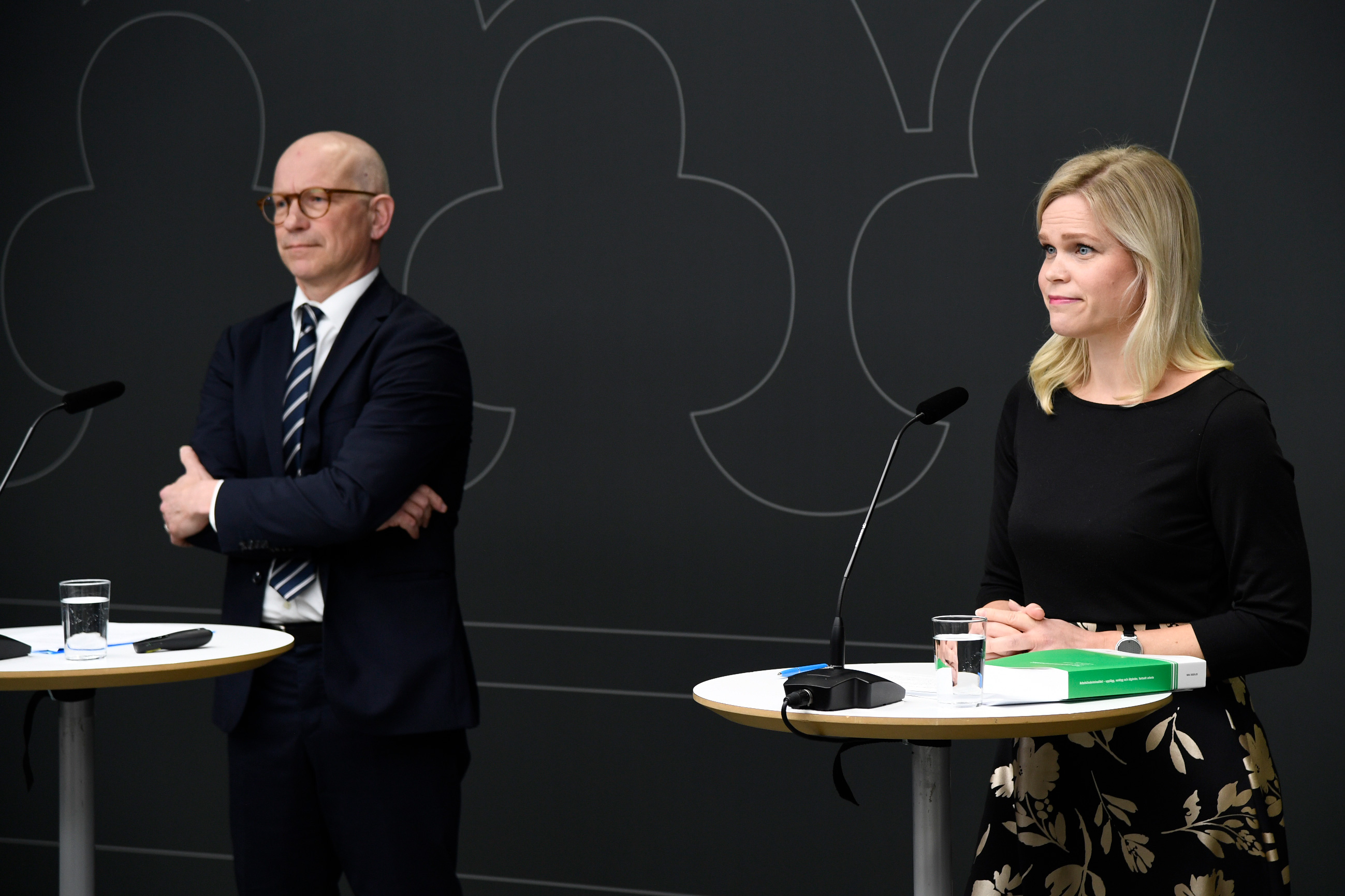 En man och en kvinna står vid podier med mikrofoner framför en mörk vägg. Mannen har på sig en kostym och kvinnan bär en svart topp. Det finns vattenglas på podierna.