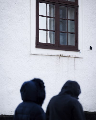 Maria och Teresa i mörka rockar och huvtröjor går förbi en vit vägg med ett fönster som har brun ram.