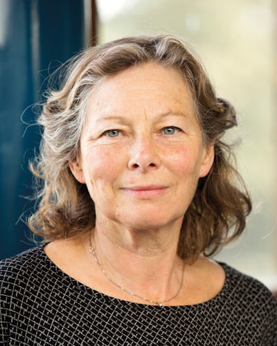 En kvinna med hår till axlarna och en mönstrad skjorta tittar rakt mot kameran. Hon står inomhus med en suddig bakgrund bakom sig.