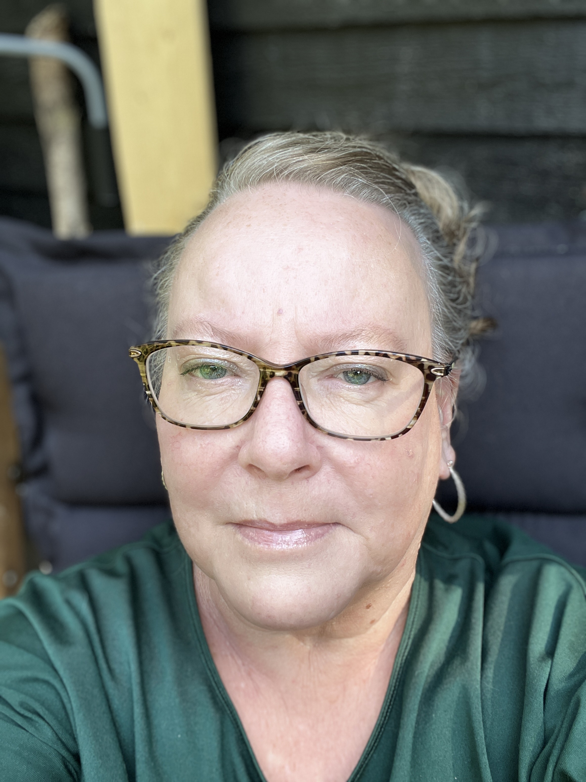 En person med glasögon och kort hår, iklädd en mörkgrön tröja, sitter utomhus med ett neutralt ansiktsuttryck.