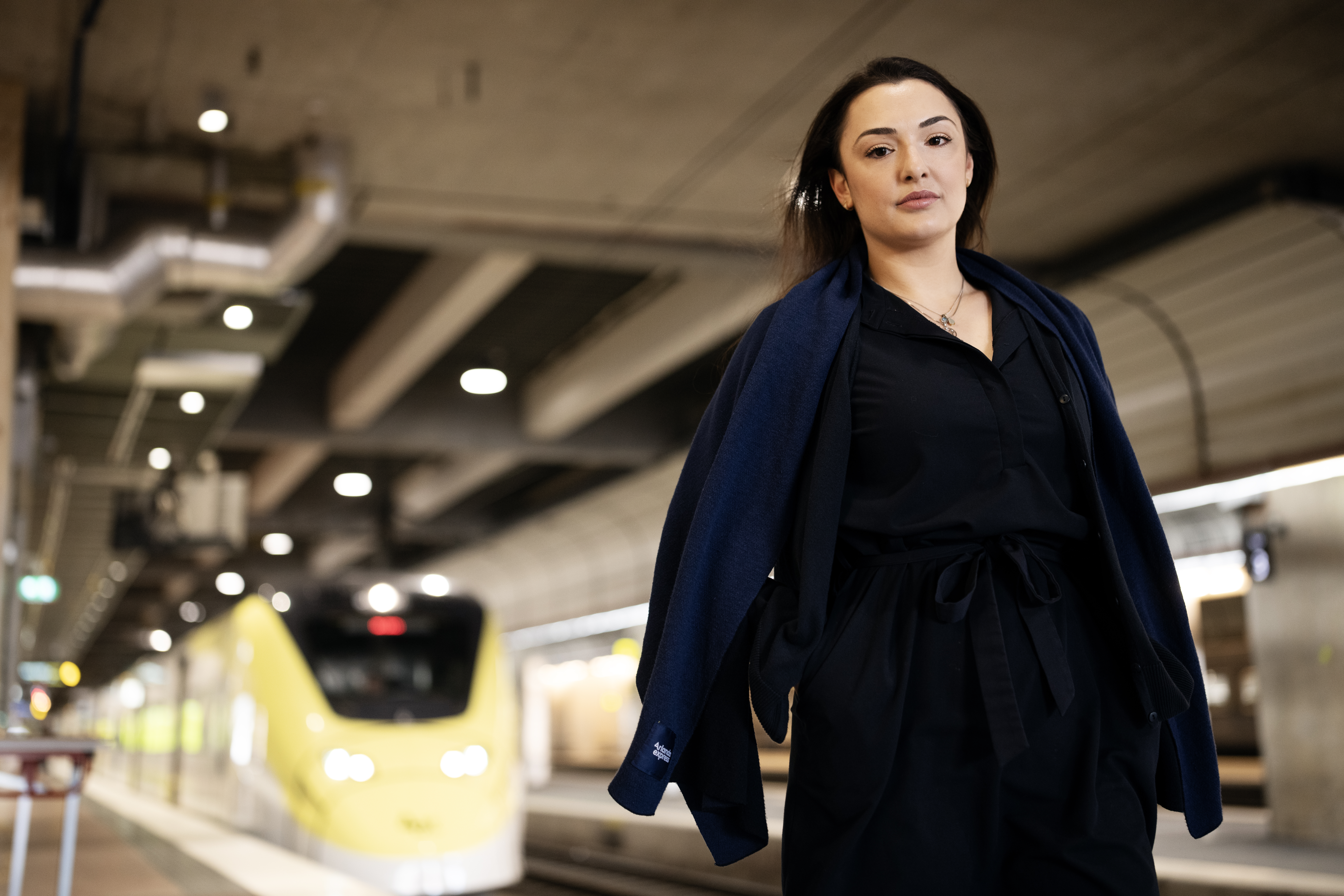 En person klädd i mörka kläder står på en tågperrong medan ett gult tåg närmar sig i bakgrunden.