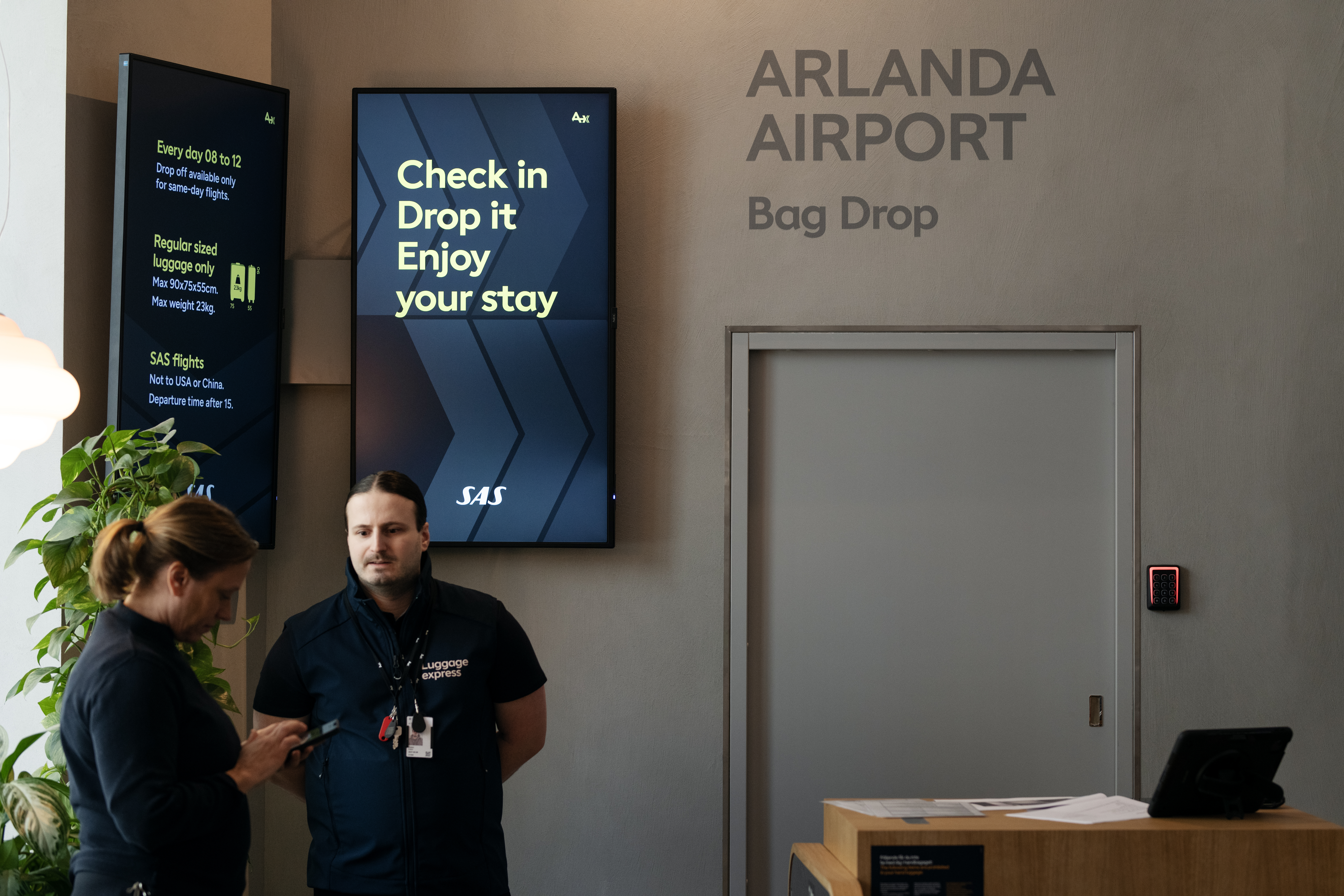 Två personer vid incheckningsområdet för bagageinlämning på Arlanda flygplats, med digitala skärmar som visar information och växt till vänster.