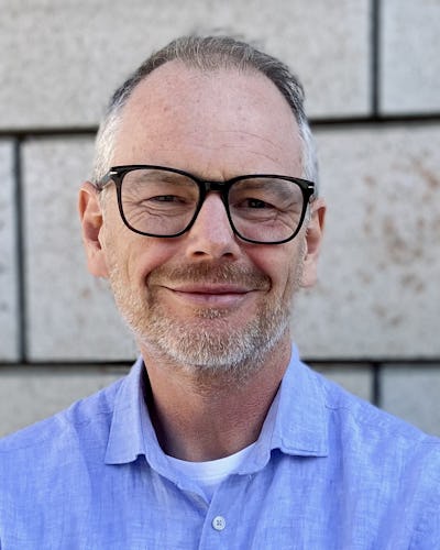Fredrik Sparring med grått hår, glasögon och kort skägg har på sig en blå skjorta och ler.