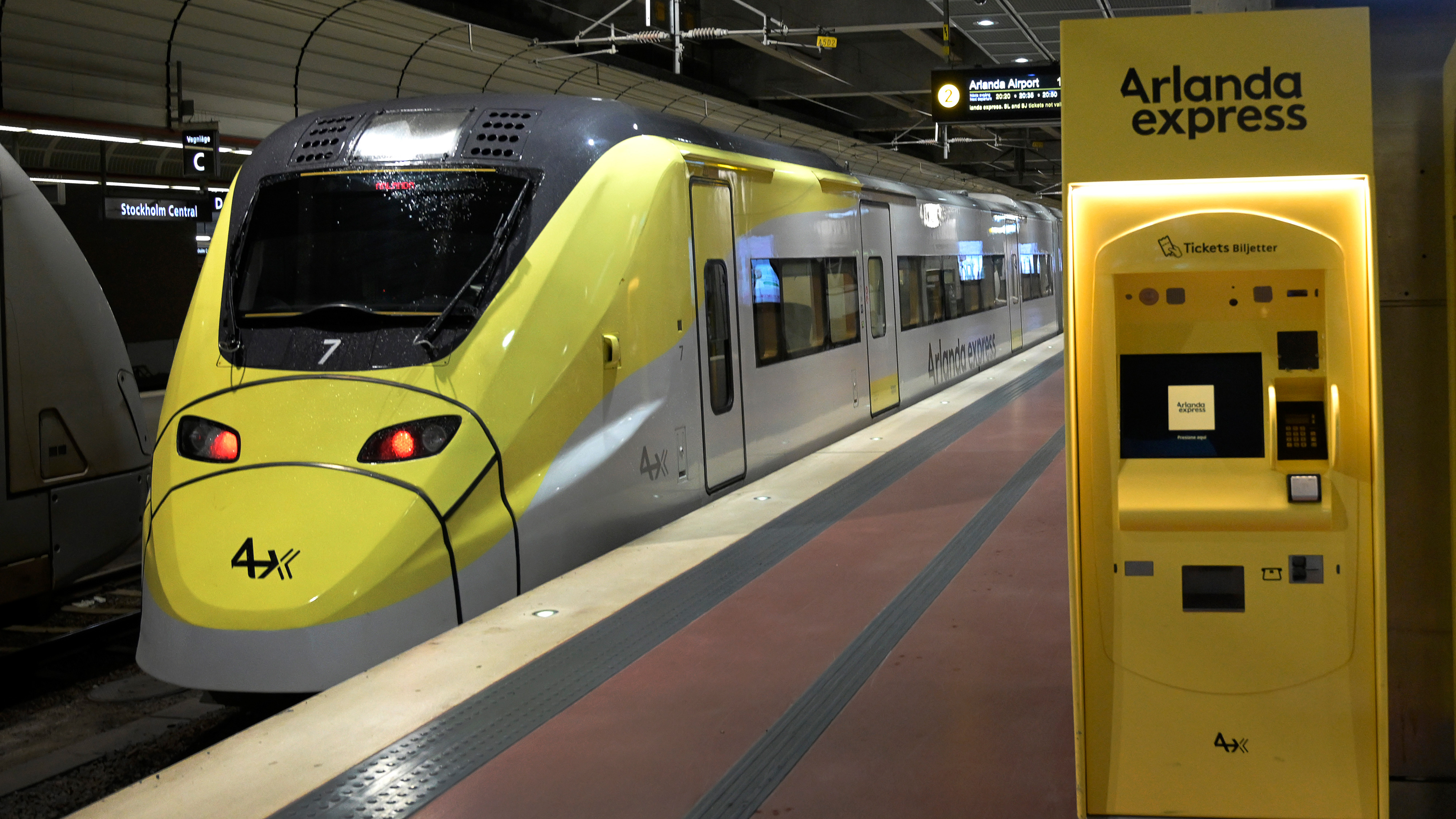 Arlanda Express-tåget i gult och grått står vid en station med en gul biljettautomat till höger.