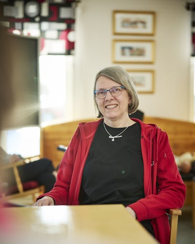 Anette Nilsson med glasögon och röd Kommunal-jacka sitter vid ett bord inomhus och tittar mot kameran. I bakgrunden finns inramade bilder och en bänk av trä.
