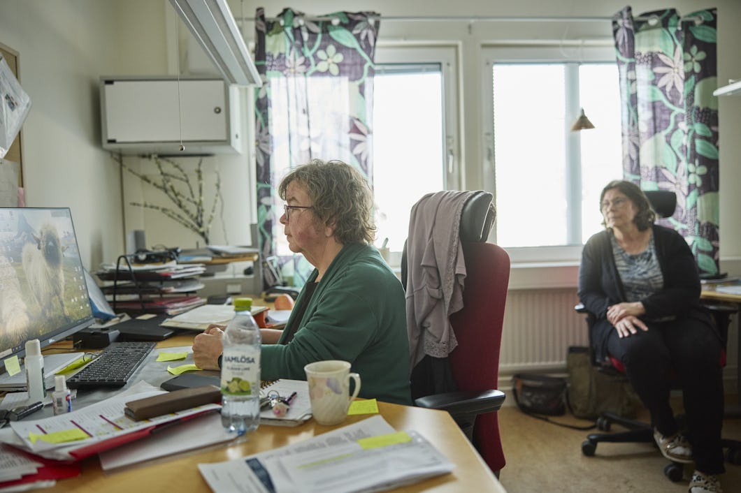 Anita Närva (i grönt) och Ingegerd Hetta (svart kofta) är på ett kontor. Anita arbetar vid ett skrivbord med en dator, Ingegerd sitter bakom henne. Det finns papper, en vattenflaska och koppar på skrivbordet. Solljus lyser i genom fönster med blommiga gardiner.