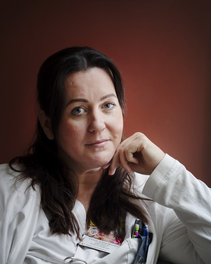 Victoria Sydäng Blomster med mörkt hår sitter med handen på hakan och har en vit rock på sig. Det finns flera pennor och en namnbricka fästa på rocken.