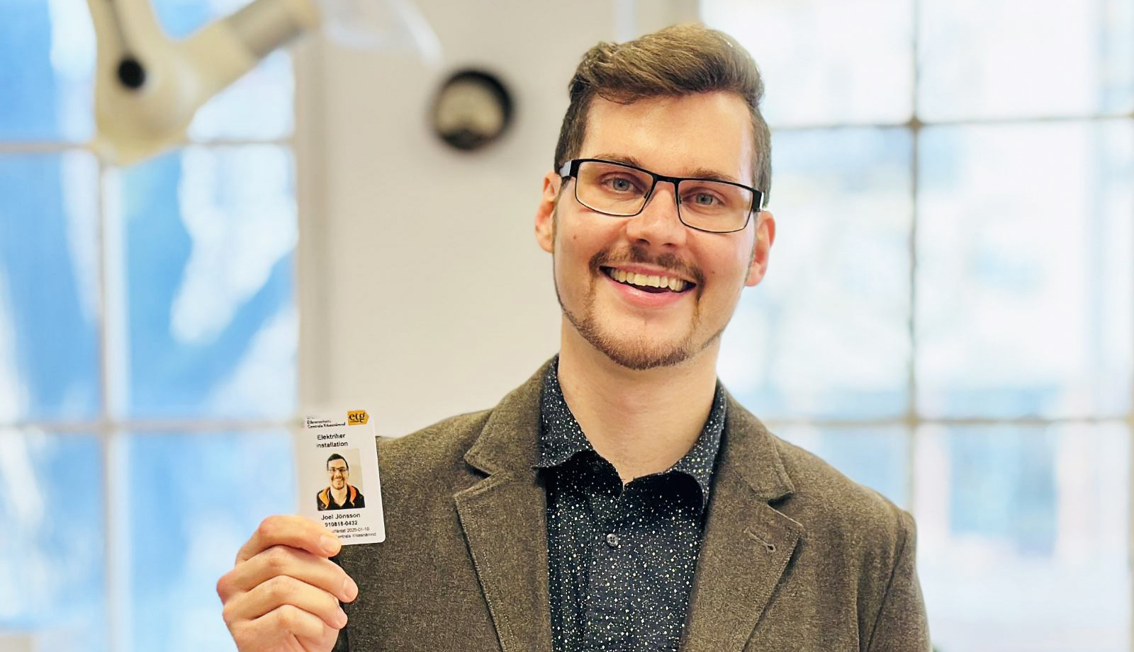 En man med glasögon ler och håller ett ID-kort med sitt foto.