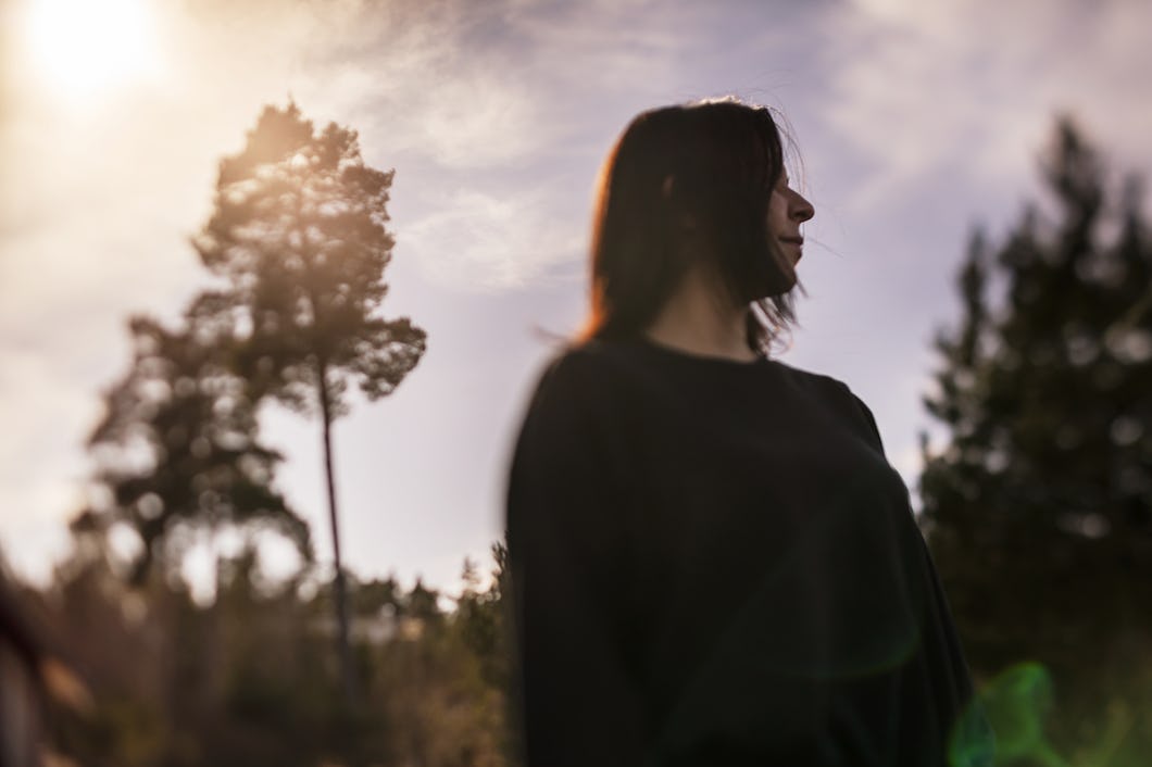 En person med långt mörkt hår står utomhus framför träd och tittar åt sidan medan solljus kommer genom himlen.