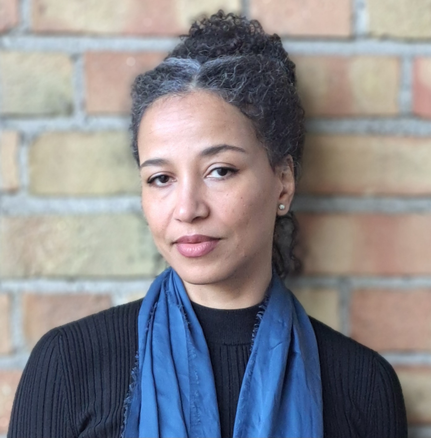 En person med lockigt hår och en blå halsduk står mot en tegelvägg och tittar direkt in i kameran.