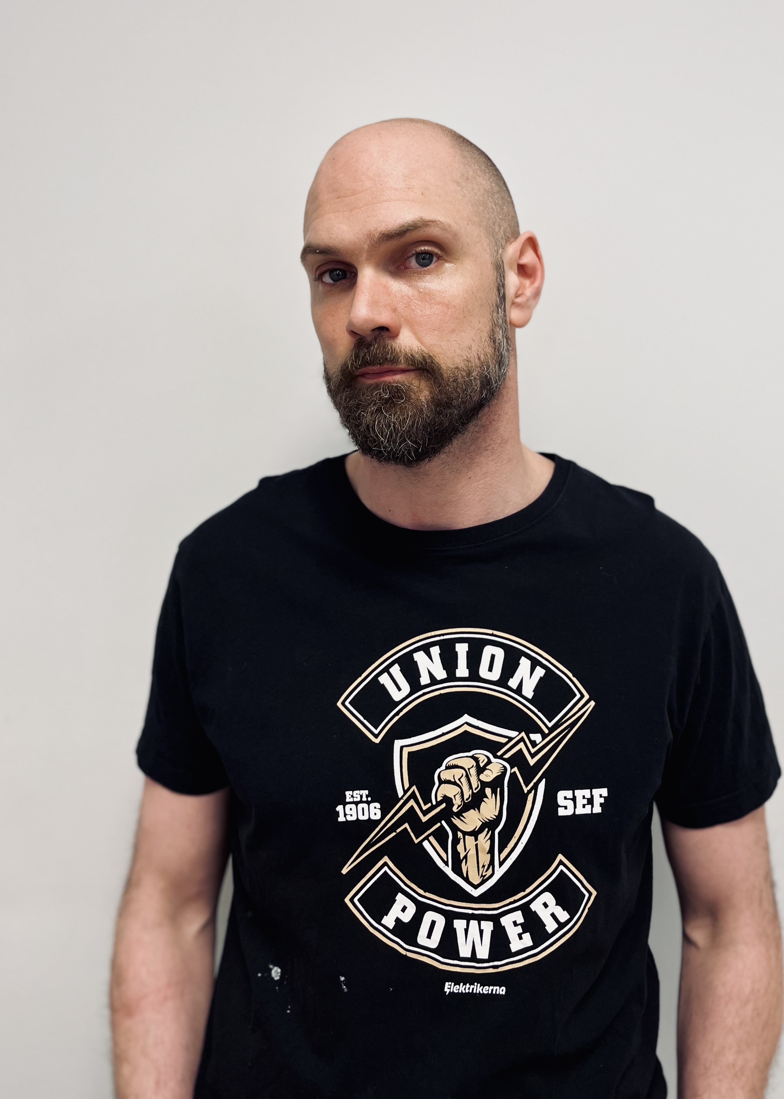 En man med skägg har på sig en svart T-shirt med texten "Union Power" och står mot en vit bakgrund.