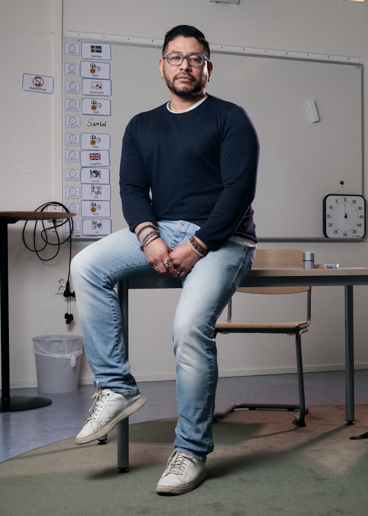 Magnus Hermansson med glasögon och vardagskläder sitter på kanten av ett skrivbord i ett klassrum. I klassrummet finns tomma stolar, en whiteboard och en rumsavdelare.