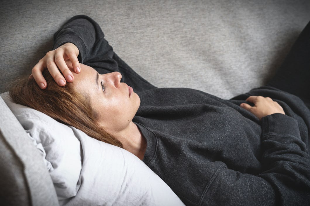 En person ligger på en soffa, vilar huvudet på en kudde och håller handen mot pannan. Personen har en mörk tröja på sig.