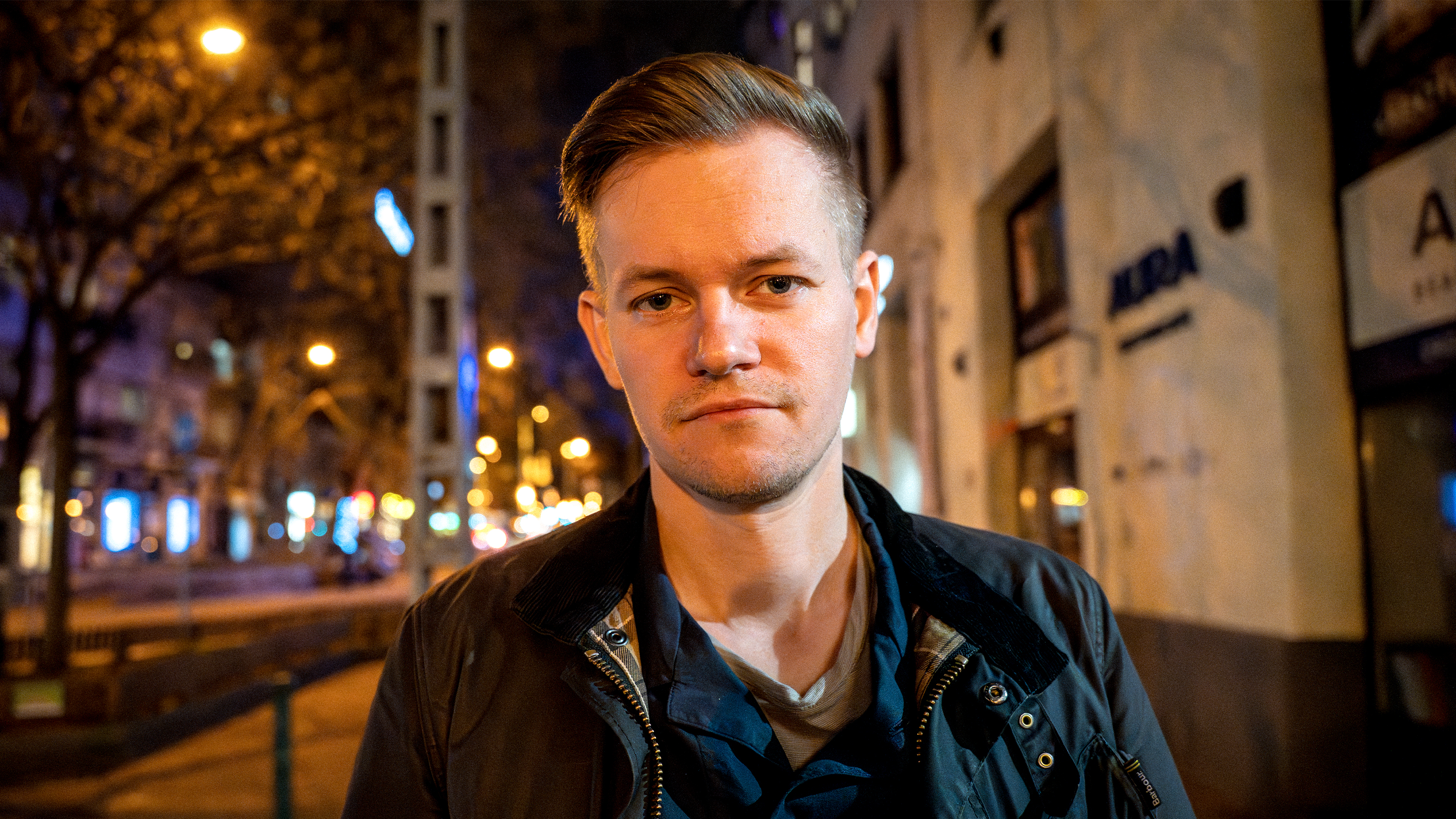 Joakim Medin står utomhus på natten på en stadsgata, med suddiga ljus och byggnader i bakgrunden.