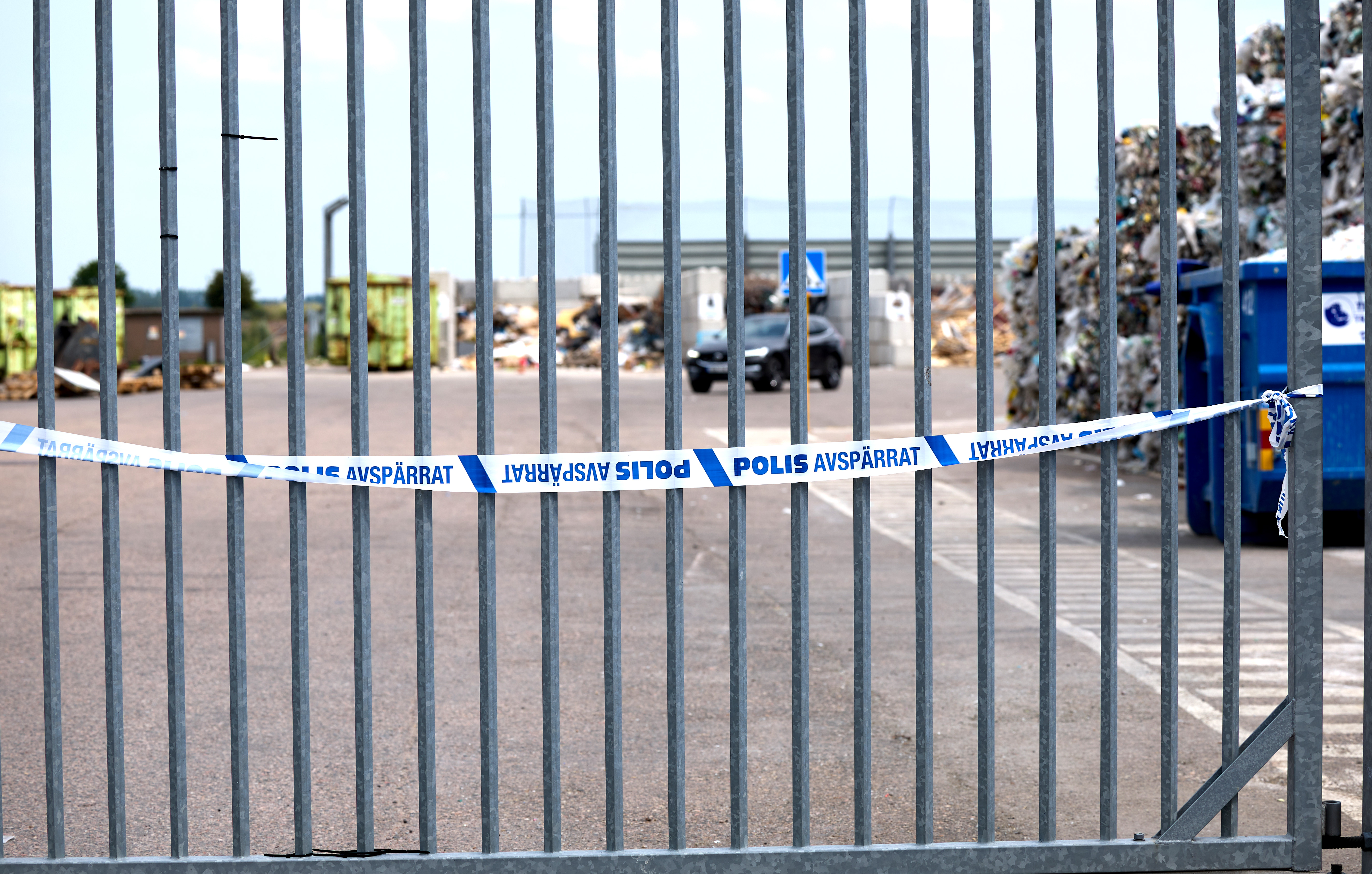 Ett avgränsat område med ett "Polis Avspärrat"-band som markerar en polisavspärrning. I bakgrunden syns staplade material och soptunnor.