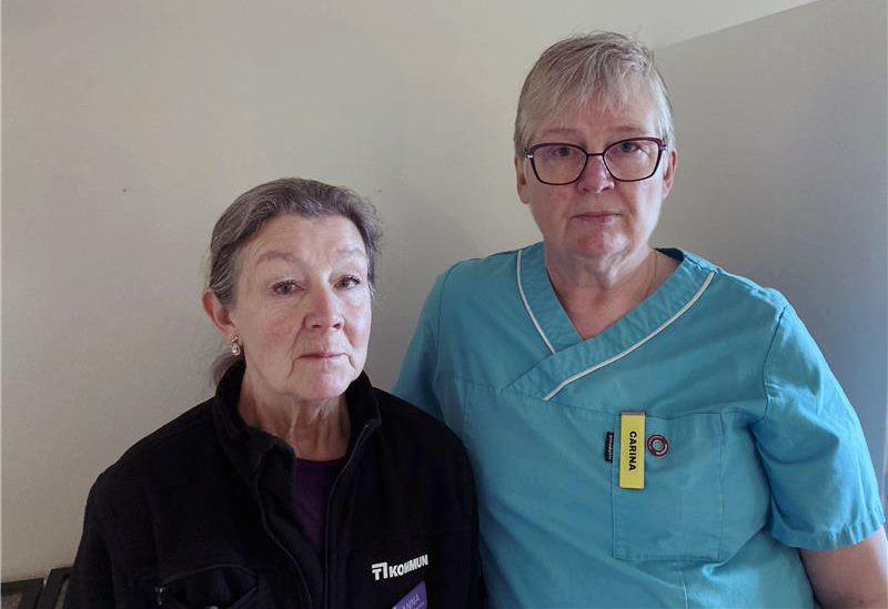 Carina Holm och Anna Gunnarsson står vända framåt. Den ena har en svart jacka på sig, den andra bär turkosa sjukhuskläder med en gul namnbricka. De står mot en neutral bakgrund.