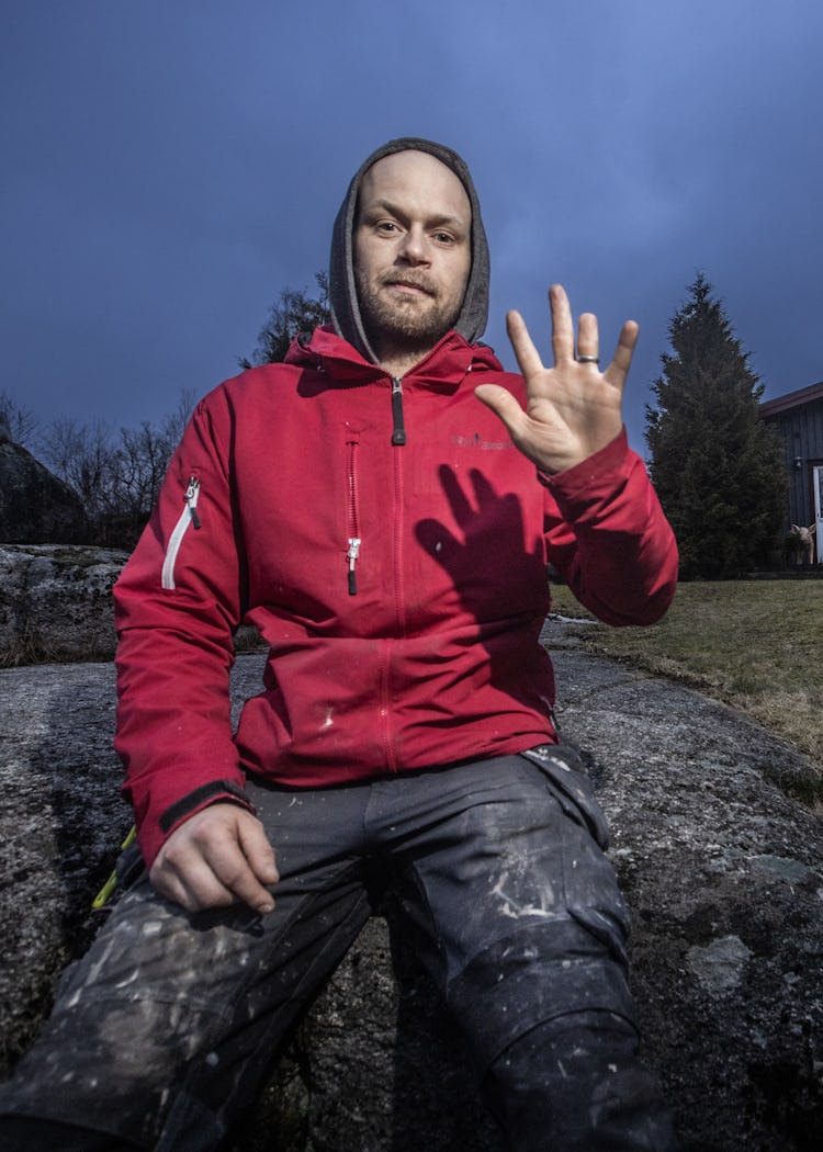 Mattias Hultgrens i en röd jacka sitter på en sten och höjer ena handen, där ringfingret är kapat. Ett hus syns i bakgrunden under en molnig kvällshimmel.