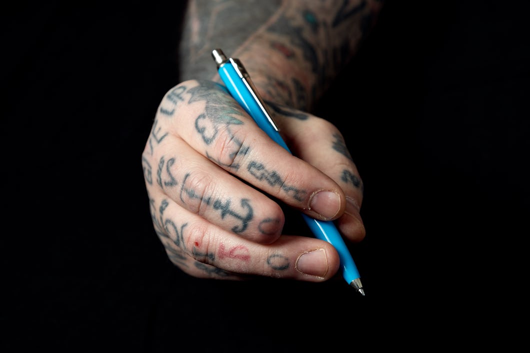 En hand med tatueringar håller i en penna mot en mörk bakgrund. Yttersta delen av ett finger är av.