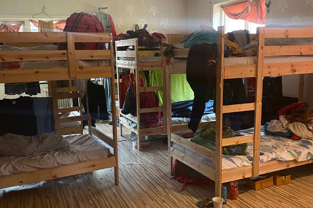 Rum med flera våningssängar av trä, kläder hänger från sängarna och området runtomkring, trägolv och fönster med gardiner.