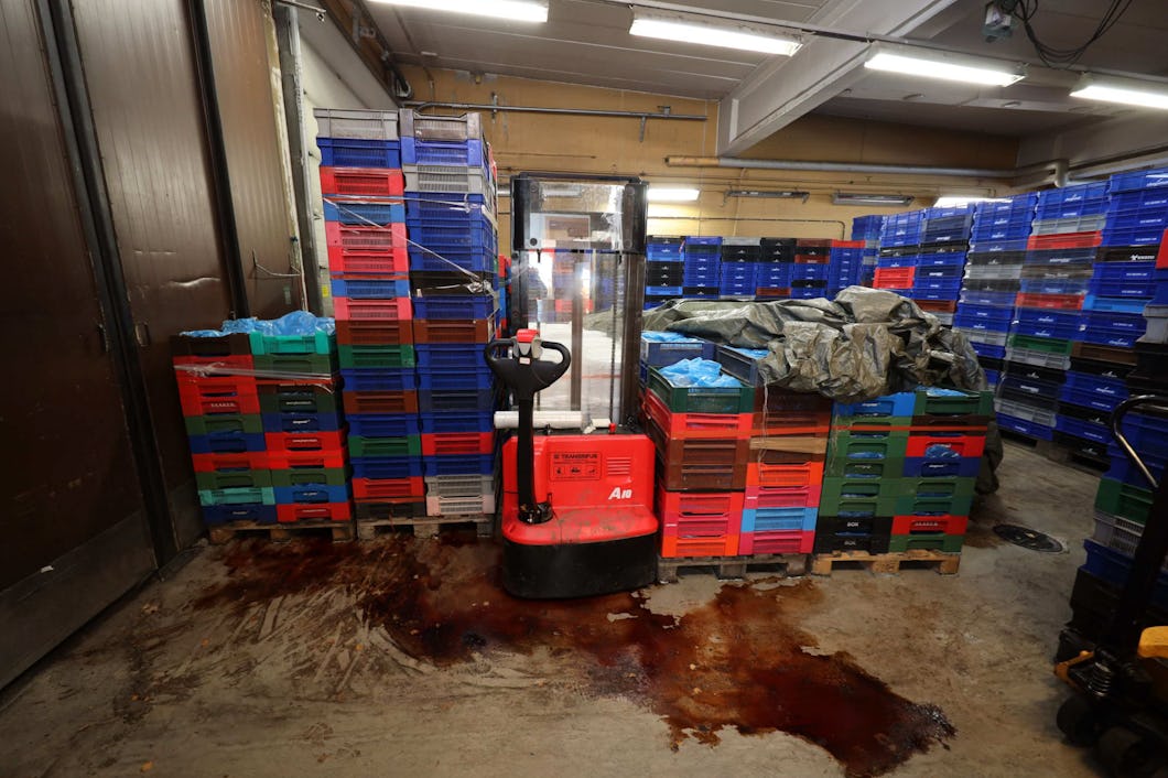 En pallyftare står parkerad i ett lagerutrymme fyllt med staplade lådor. En presenning täcker några av lådorna, och vätska har spillts på golvet.
