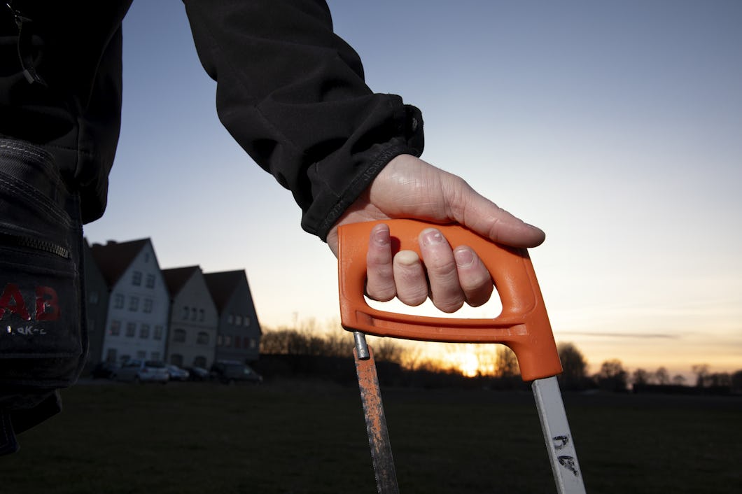 En hand som håller i en handsåg med en orangefärgad handtag mot en bakgrund av solnedgång, med byggnader längre bort.