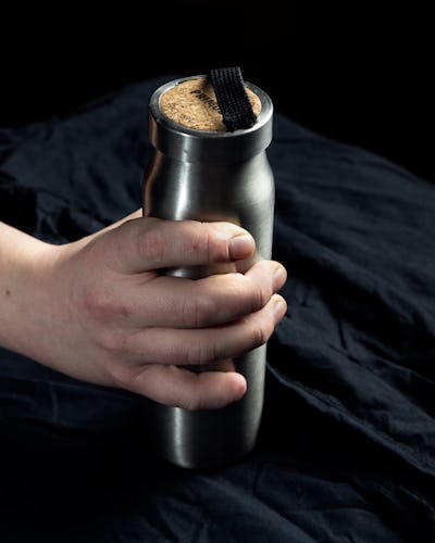 En hand håller i en vattenflaska av rostfritt stål med en korktopp och ett svart band, mot en mörk tygbakgrund.