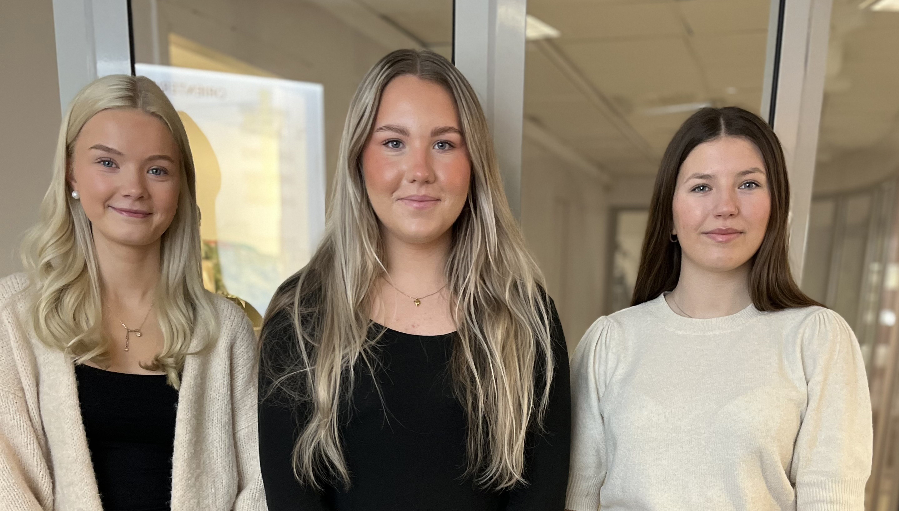 Tre unga kvinnor står inomhus, vända framåt.