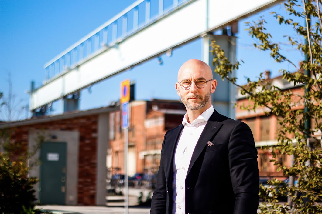 En skallig man i kostym står utomhus framför industribyggnader och metallstrukturer en soldag.