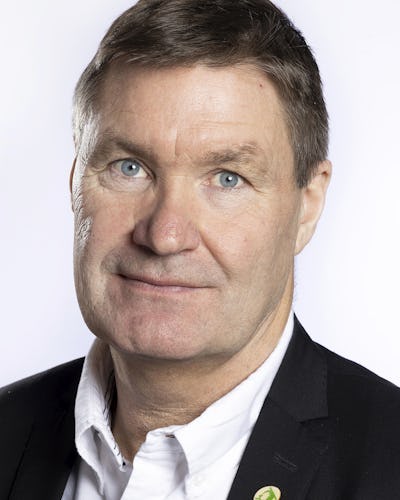 Andreas Karlsson med kort brunt hår har en vit skjorta och en mörk kavaj. Han står framför en ljus enfärgad bakgrund.