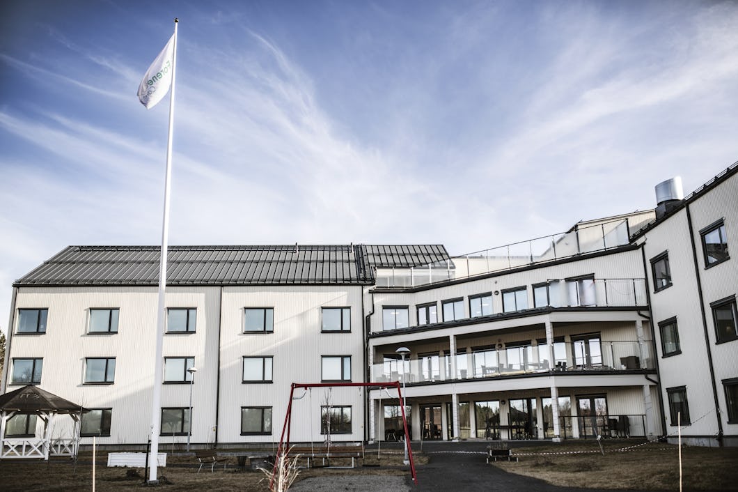En byggnad med flera våningar och stora fönster. Det finns en flaggstång och ett gungställ i trädgården framför huset.
