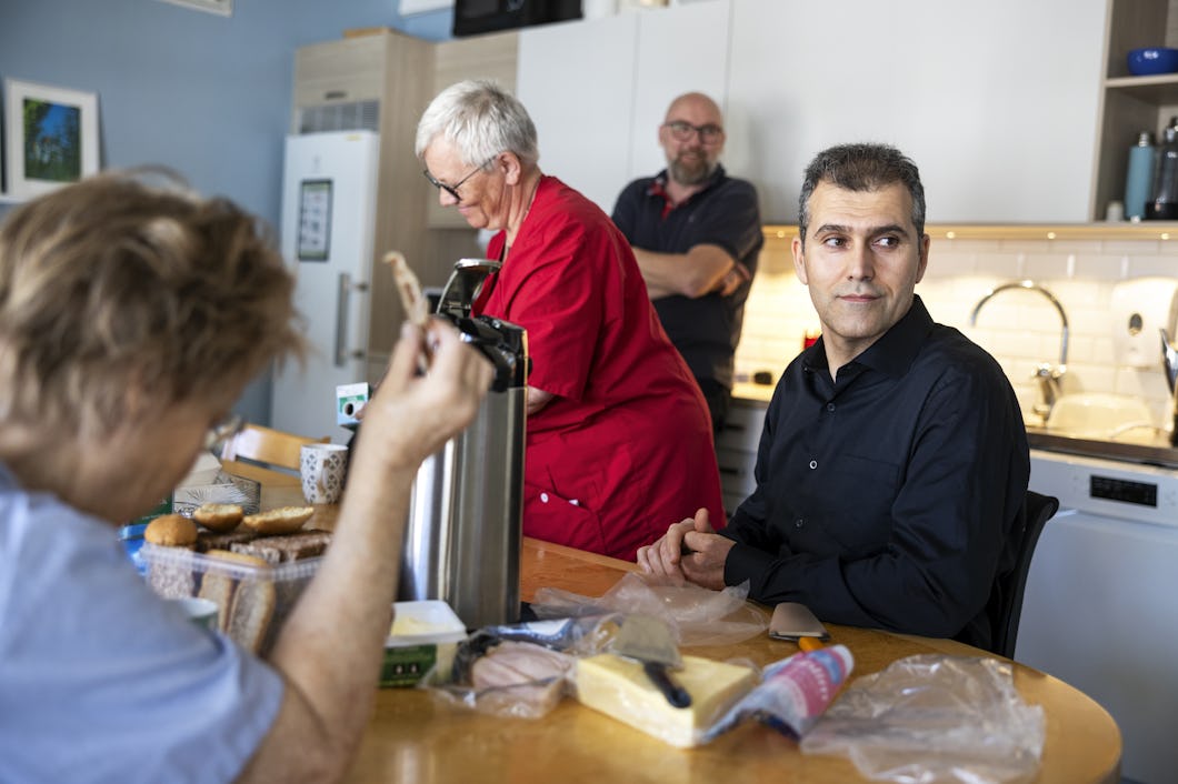 Zana Rafaat och tre andra personer är i ett kök runt ett bord med bröd, ost och annan mat. En kvinna har på sig ett rött förkläde och lagar mat, medan de andra sitter eller står nära.