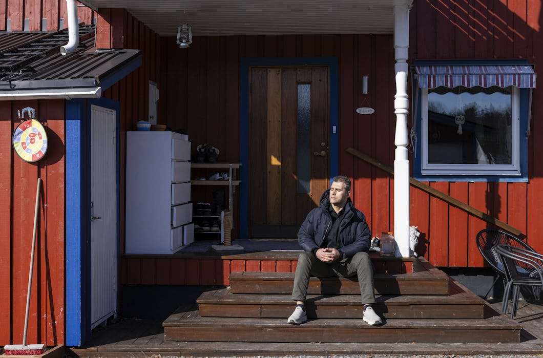 Zana Rafat sitter på trappan utanför ett rött trähus med vita kanter och tittar lite åt sidan i dagsljus.