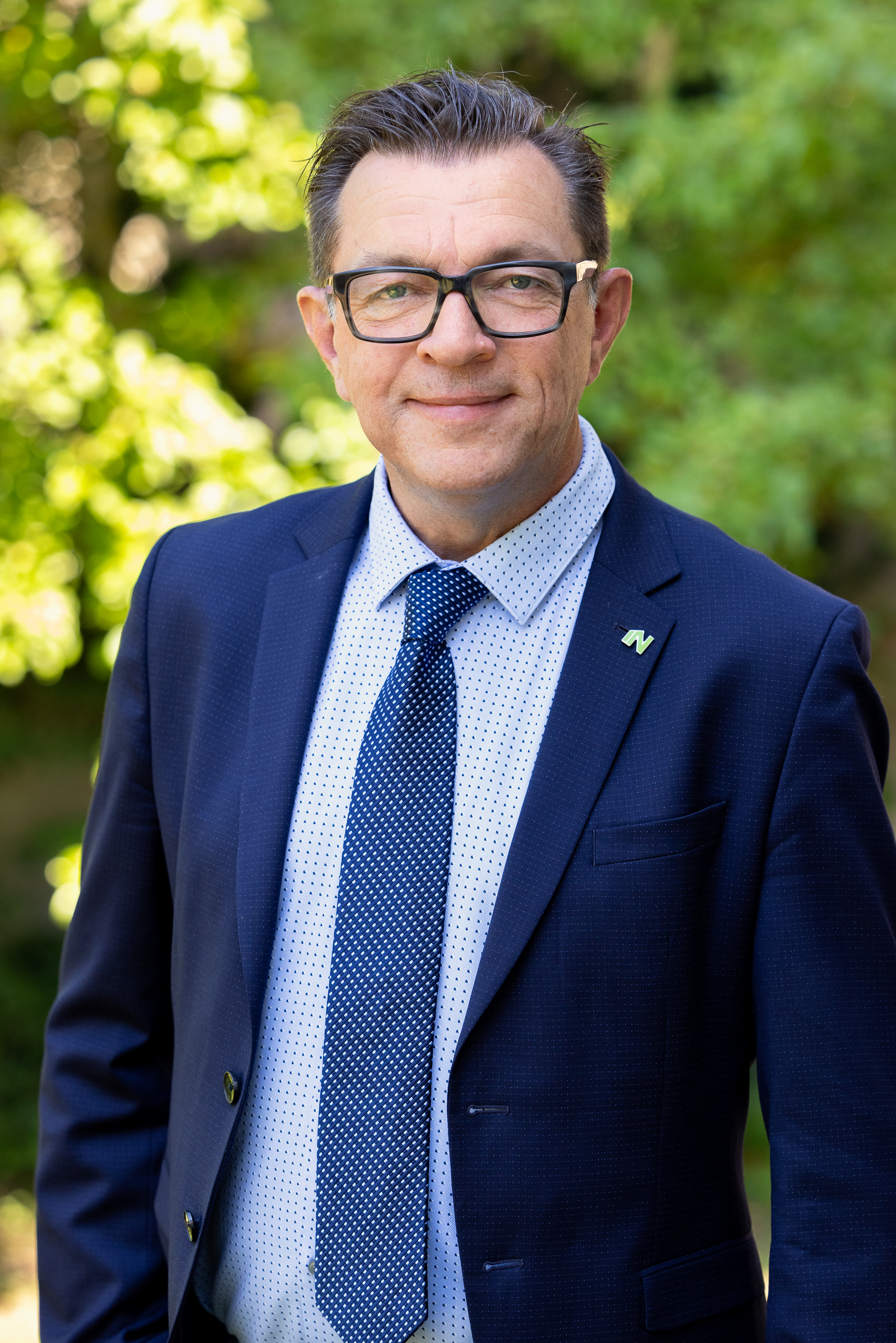 En man i kostym och slips står utomhus framför växtlighet och tittar mot kameran med en svagt leende.