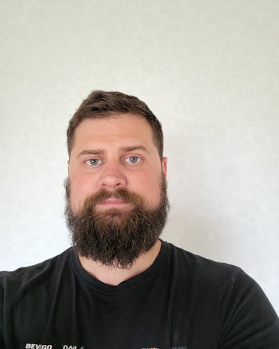 En man med skägg, kort brunt hår och blå ögon har på sig en svart T-shirt och är vänd mot kameran. Bakgrunden är ljus och enkel.