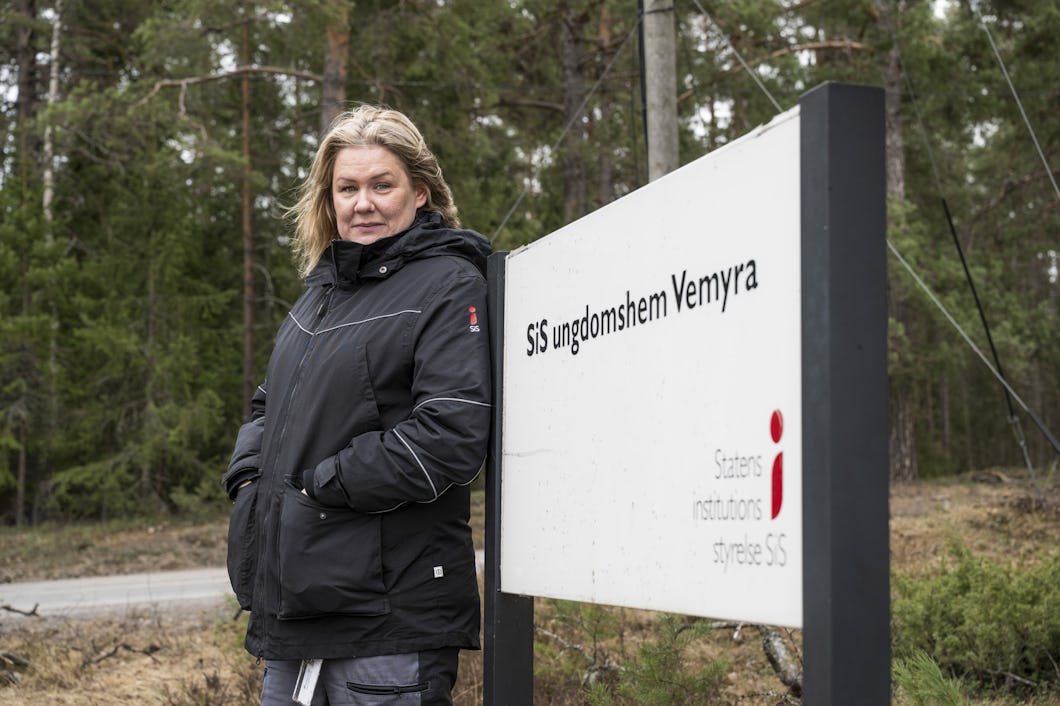 Jennie Salomonsson i svart jacka står bredvid en skylt där det står "SiS ungdomshem Vemyra" i en skogsmiljö utomhus.