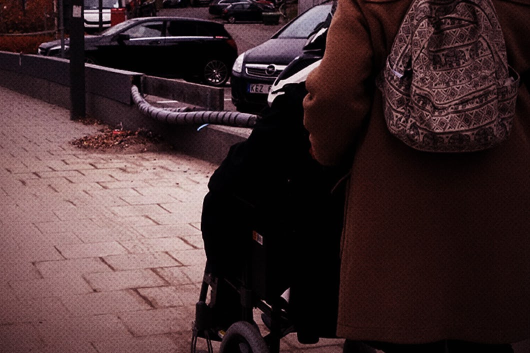 En person med en ryggsäck med mönster går bredvid någon som sitter i rullstol på en trottoar i staden nära parkerade bilar.