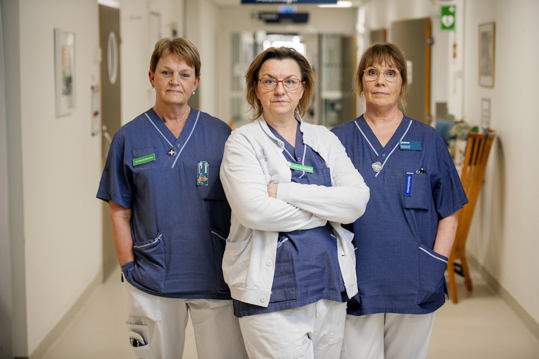 Birgitta Tocklin, Karmena Jukic och Suzanne Ask i sjukvårdskläder står i en sjukhuskorridor och tittar mot kameran. Kvinnan i mitten har armarna i kors, medan de andra har händerna vid sidorna.