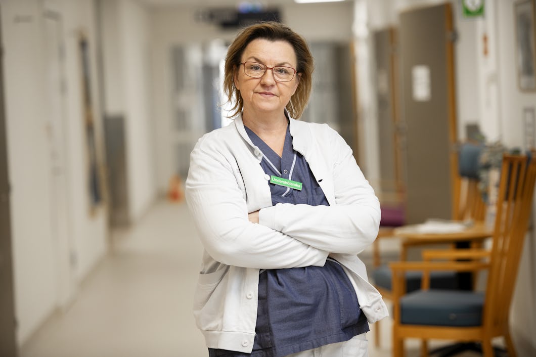 Karmena Jukic i medicinska arbetskläder och vit rock står med armarna i kors i en sjukhuskorridor och tittar mot kameran.