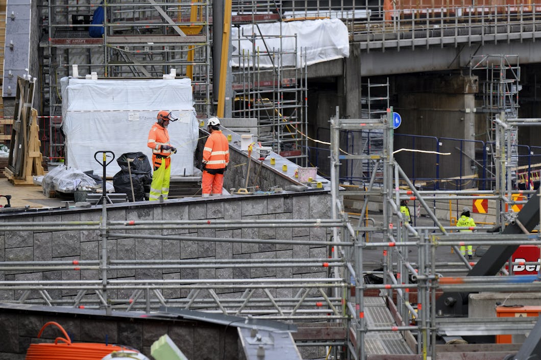 Två byggarbetare med skyddsutrustning står på en byggarbetsplats omgivna av ställningar, metallbalkar och byggmaterial.