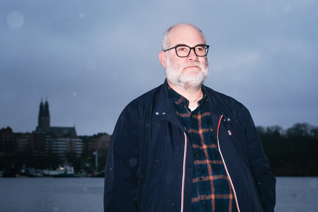 En man med glasögon och skägg står utomhus utomhus en stadsmiljö med vatten och byggnader under en molnig himmel.