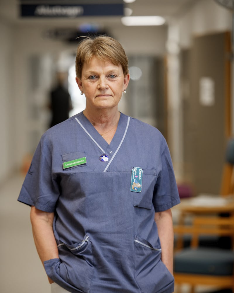 Birgitta Tocklin i sjukhuskläder står i en sjukhuskorridor med händerna i fickorna och tittar rakt mot kameran.