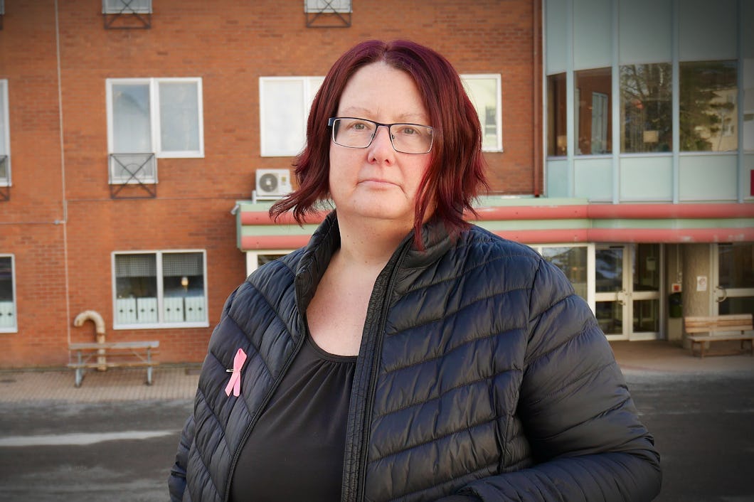 Camilla Munk Utter med axellångt rött hår och glasögon står utanför en byggnad av tegel. Hon bär en svart jacka med ett rosa band som brosch.