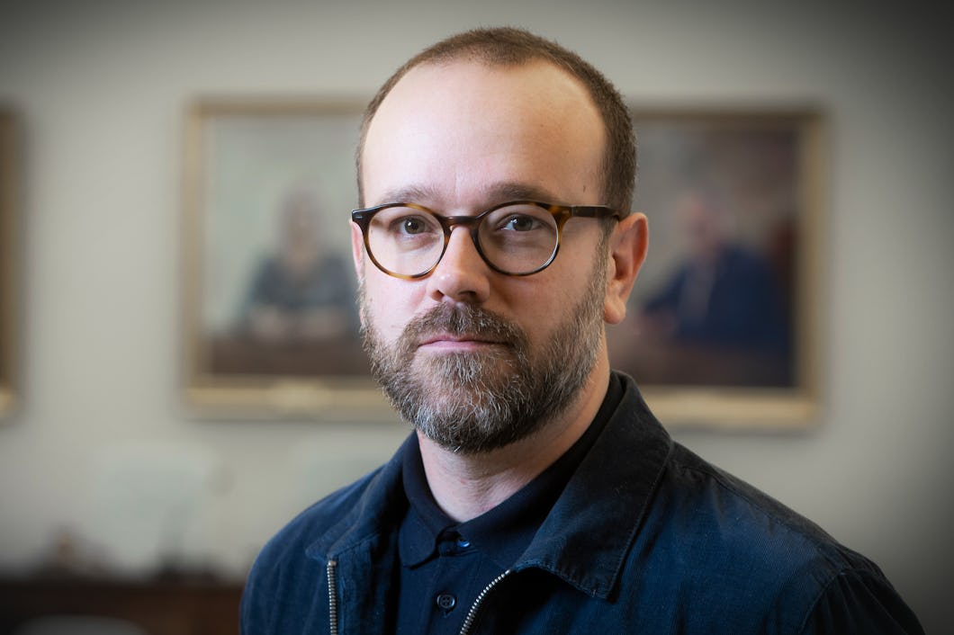 Erik Nises med glasögon och skägg står inomhus, iförd en jacka, med suddiga porträtt i bakgrunden.