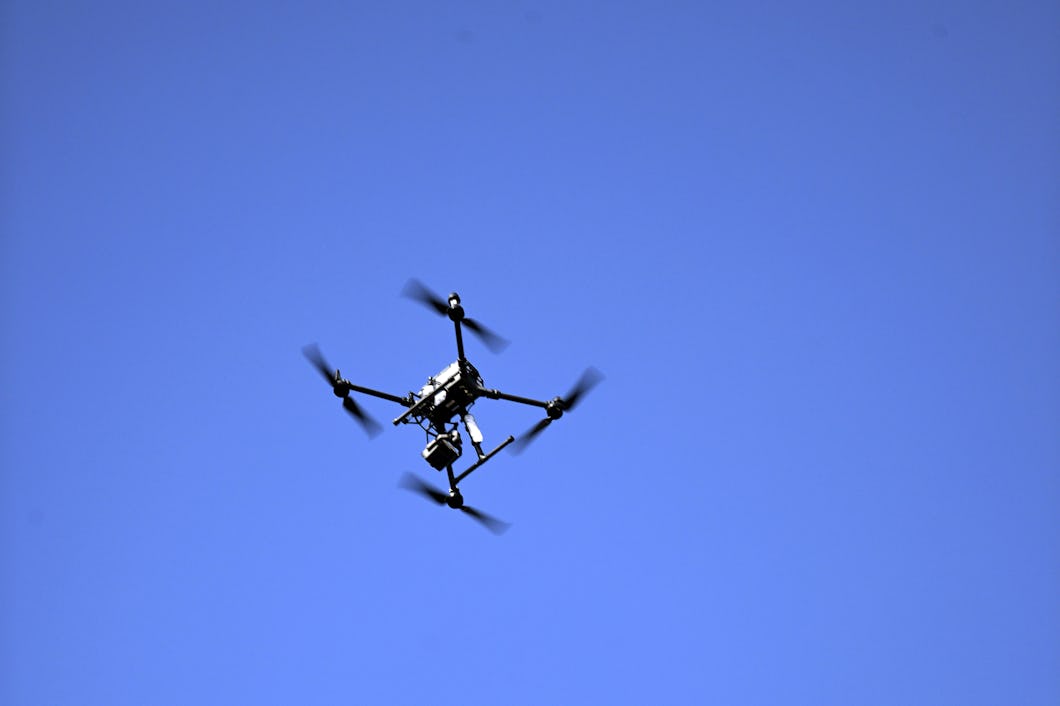 En drönare med fyra rotorer och en monterad kamera flyger mot en molnfri himmel.