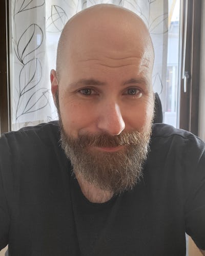 En man med skägg sitter inomhus framför ett fönster med tunna gardiner som har ett bladmotiv.